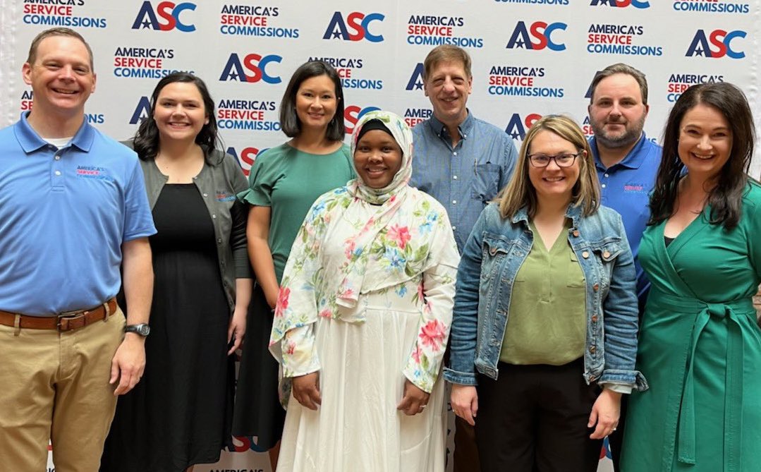 ASC Team photo. 8 staff members pose together in front of ASC step and repeat backdrop.